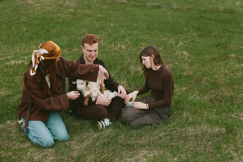 Relaxed outdoor scene with friends and family sharing a gentle moment with a goat kid on a grassy field, as they pet and interact, capturing a warm rural lifestyle moment of connection and care.