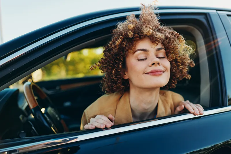 A woman with curly hair leans out of a car window, eyes closed, smiling gently as the breeze touches her face. A candid lifestyle moment with retro film color tones, conveying relaxed mood, freedom,