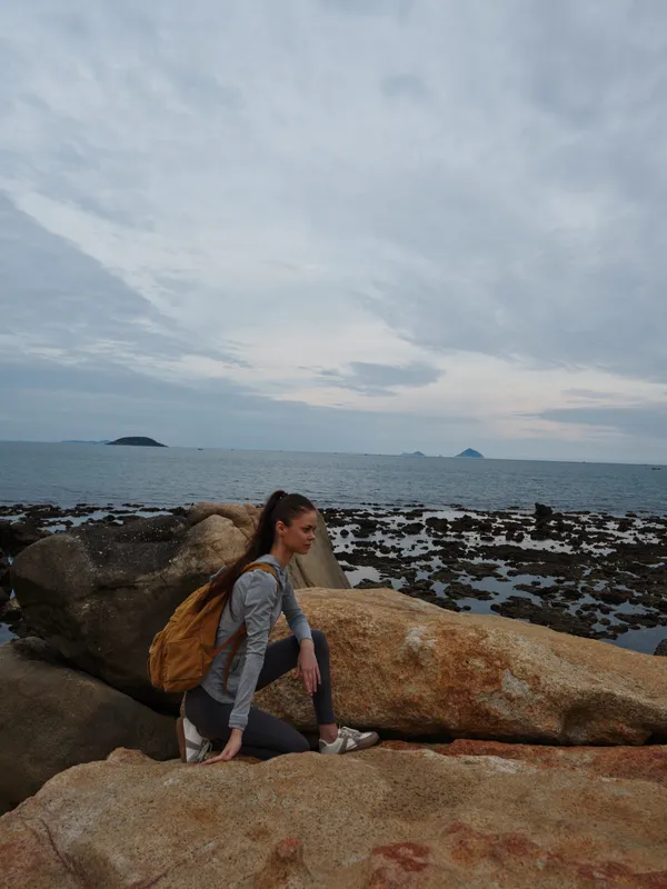 Serene woman sitting on rocky shore, wearing casual outfit and backpack, enjoying coastal view and cloudy sky