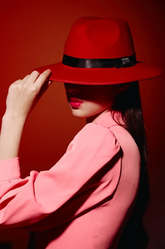 Fashionable woman in pink outfit wears striking red hat covering eyes against vivid red background. Vibrant colors emphasize bold makeup and confident stylish pose.