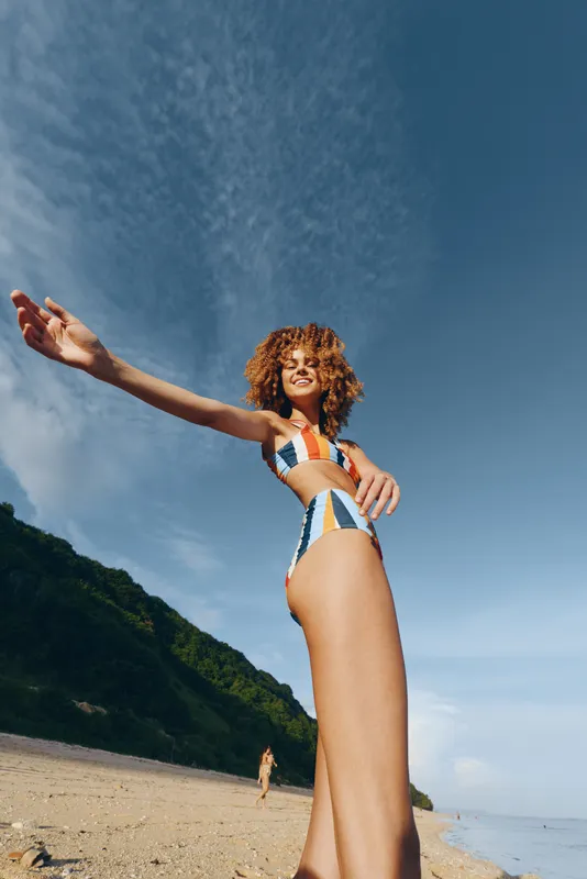 Woman in striped swimsuit on sandy beach, arms raised toward blue sky, curly hair in the wind, sunny summer day, ocean horizon and green hills behind, relaxed pose, natural light