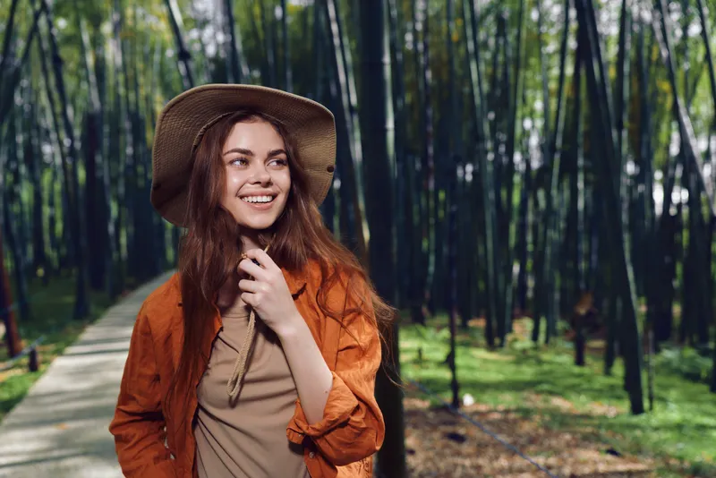 Woman with hat smiling on a forest path, outdoors portrait of a happy young adult in casual jacket enjoying a sunlit nature walk among tall trees and green scenery.