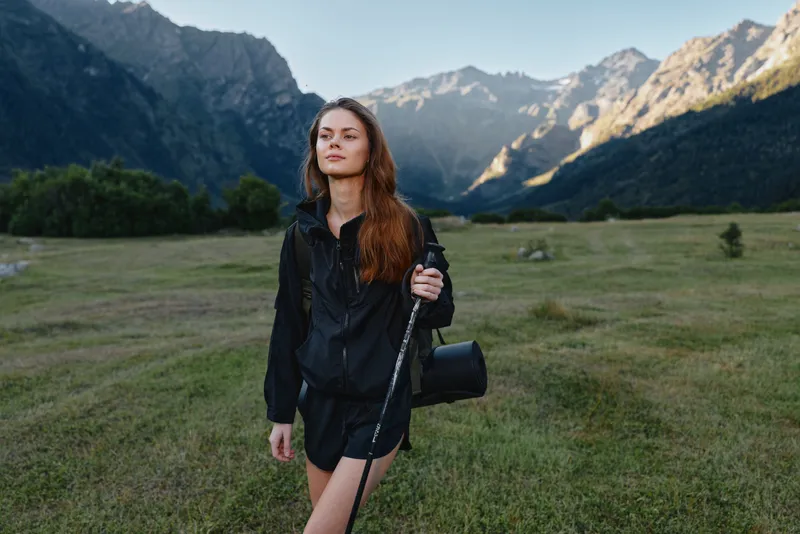A solo woman stands in a wide mountain meadow, wearing a black rain jacket and shorts, holding a trekking staff, and gazing into the distance with a confident, adventurous expression.