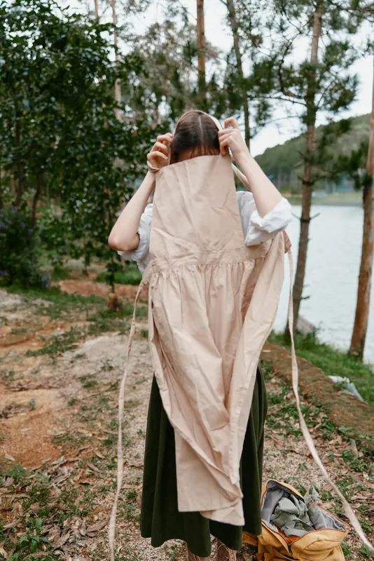 A woman in a countryside setting putting on a beige apron, showcasing a blend of nature and home cooking, perfect for promoting healthy living and culinary arts