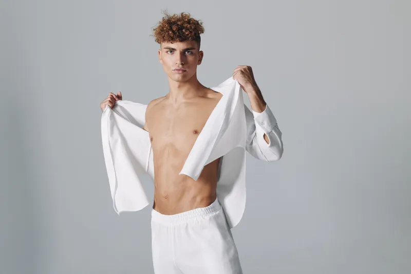 Young man with curly hair posing shirtless while holding open a white shirt, wearing white pants against a plain gray background in studio. Male model with confident expression and athletic build.