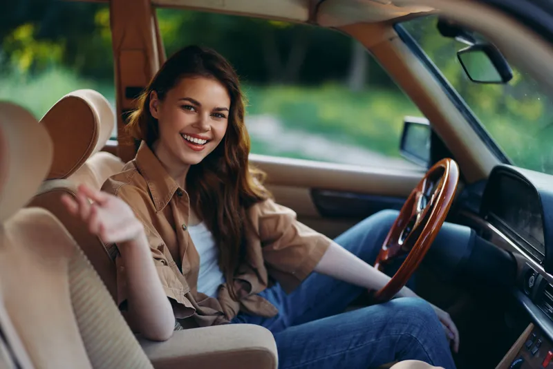 Cheerful young woman in casual outfit sitting in vintage car, enjoying a sunny day outdoors in nature, conveying happiness and freedom.