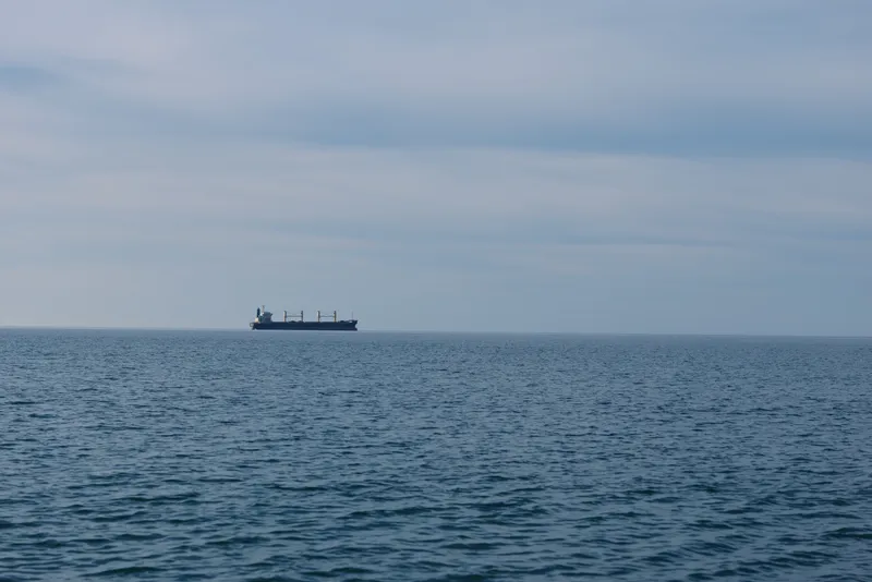 Cargo ship sailing on a calm sea with distant horizon and blue sky, a quiet maritime scene featuring a lone vessel at sea and expansive water.