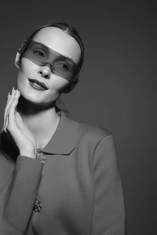 Young white woman with pale skin wears oversized transparent sunglasses and knitted sweater, posing with hand near face in studio. Concept of modern fashion and portrait photography.