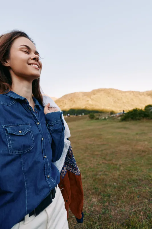 Woman smile outdoors in grassy field wearing denim shirt and carrying jacket over shoulder, portrait of happy person enjoying nature with clear sky, distant hills and warm sunlight.