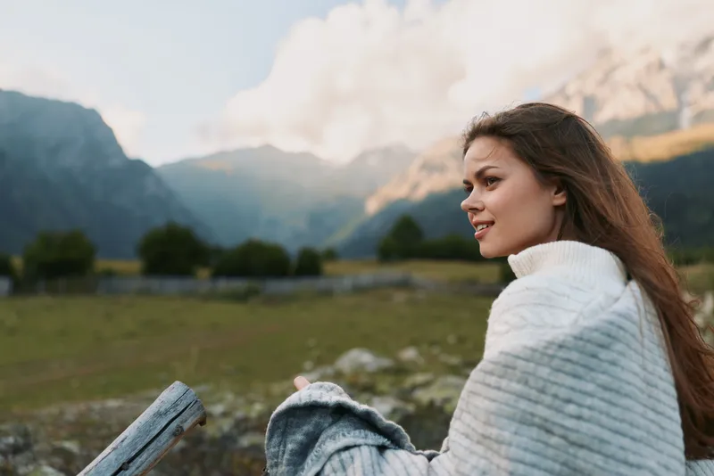 Young woman wrapped in a white shawl sits beside a wooden fence, smiling softly as majestic mountains rise in the distance on a bright, clear day.