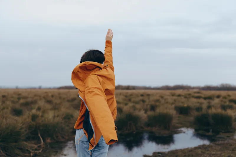 Woman outdoor lifestyle in orange jacket walking along a river path, turning face to sky, casual smile, soft evening light, open countryside, calm wind, natural colors, spring breeze