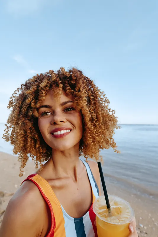 Smiling young woman with curly hair holds a cold drink with a straw on the beach under clear blue sky, enjoying sunny summer day near calm sea water.