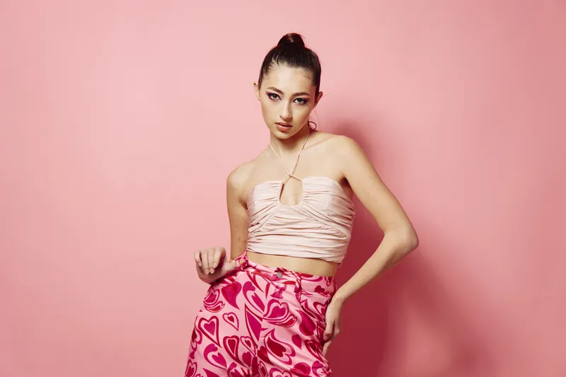 Stylish young woman poses confidently wearing creative pink patterned pants and a textured cropped top against a soft pink backdrop. Fashion and beauty concept with expressive attitude.