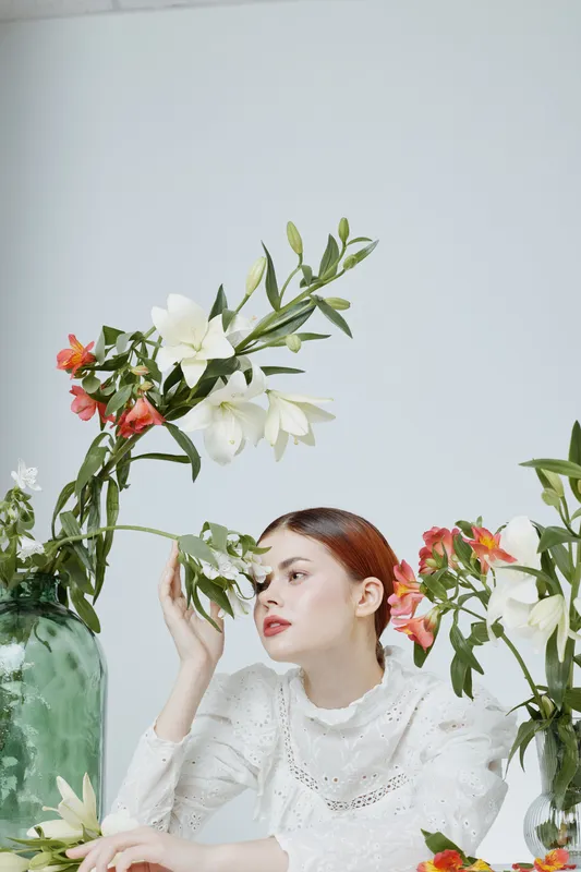 Beautiful young woman with red hair in white dress surrounded by colorful flowers in a minimalistic studio setting, expressing calmness and elegance