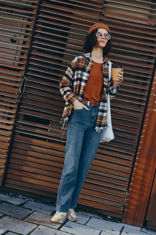 Woman with coffee on street in casual autumn outfit, sunglasses and beanie, totebag and plaid shirt conveying candid lifestyle, authenticity and warm golden hour glow mood.