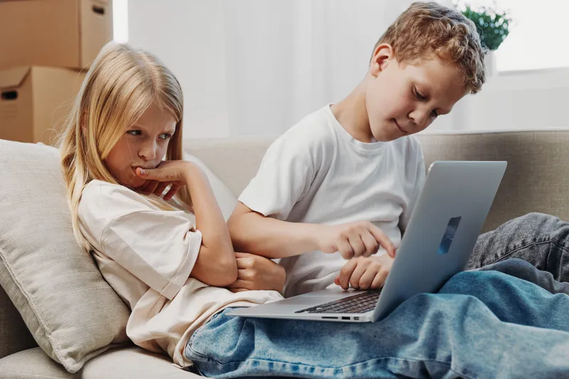 Two children sitting on a couch using a laptop, a boy focused on the screen and a girl looking thoughtful. Concept of childhood, technology, sibling interaction, and learning at home.