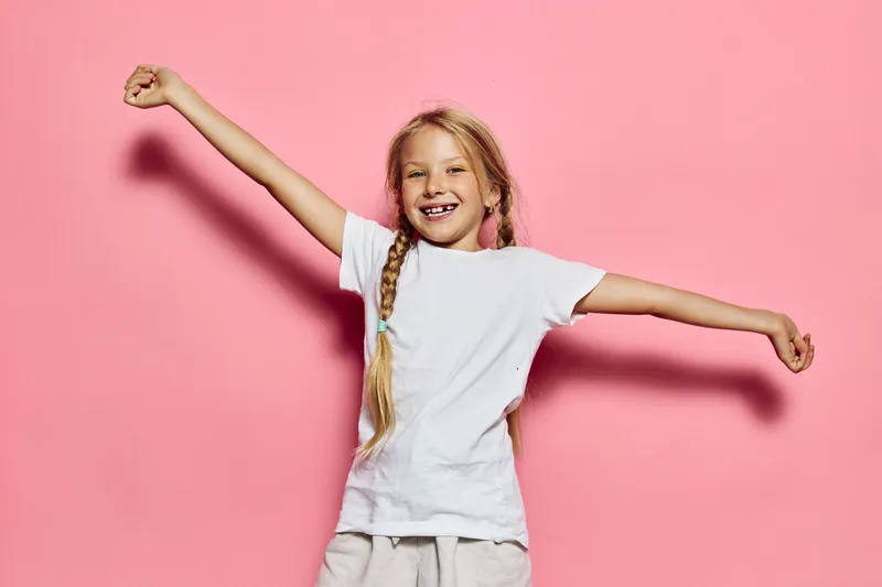 Happy child with blonde hair in braids wearing white t-shirt raising arms in celebration against pink background. Studio shot with bright soft lighting and joyful expression.