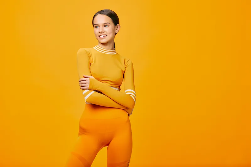Woman in athletic orange outfit stands against a vibrant orange backdrop, showcasing fitness and sport lifestyle with confident pose and energetic presence.