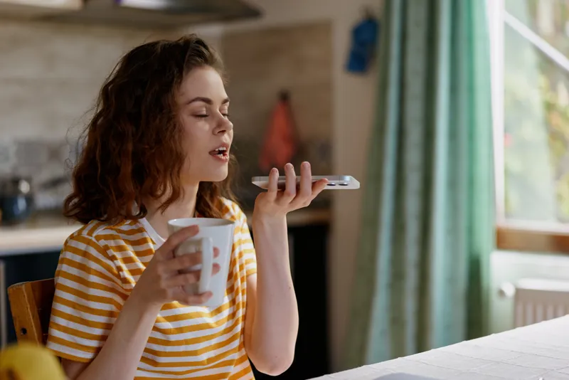 Cheerful woman at home, sipping coffee from a mug while talking on a phone, casual striped shirt, cozy morning atmosphere, natural light, relaxed lifestyle moment