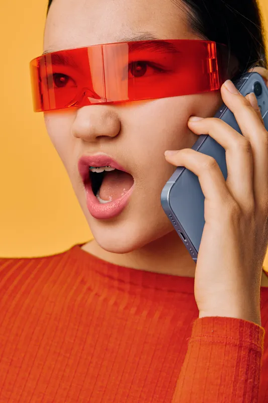 Woman with futuristic red glasses talking on smartphone with open mouth against orange background. Female model wearing modern eyewear and orange shirt in closeup portrait for technology and