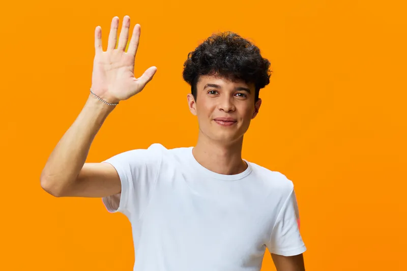 young man waving hand with cheerful smile, curly hair, wearing white t-shirt against vibrant orange background, friendly gesture in casual style