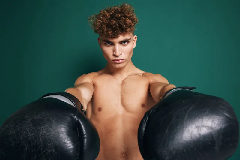 Young muscular man with curly hair wearing black boxing gloves in fighting stance against green background. Fitness, training and sport concept with focused athlete.