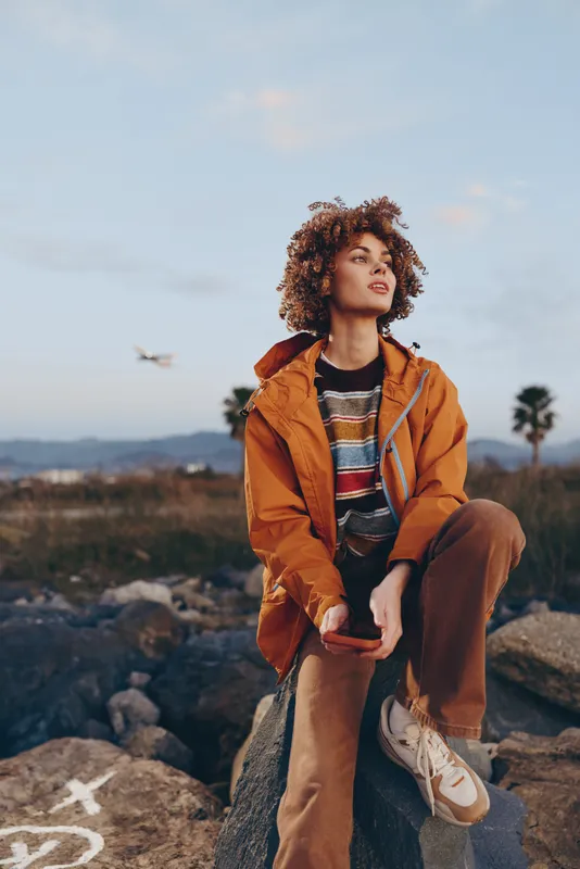 woman lifestyle outdoors portrait curly jacket travel nature sky adventure, young woman with curly hair in an orange jacket sits on rocks, daylight mountains landscape and clouds fill the sky,