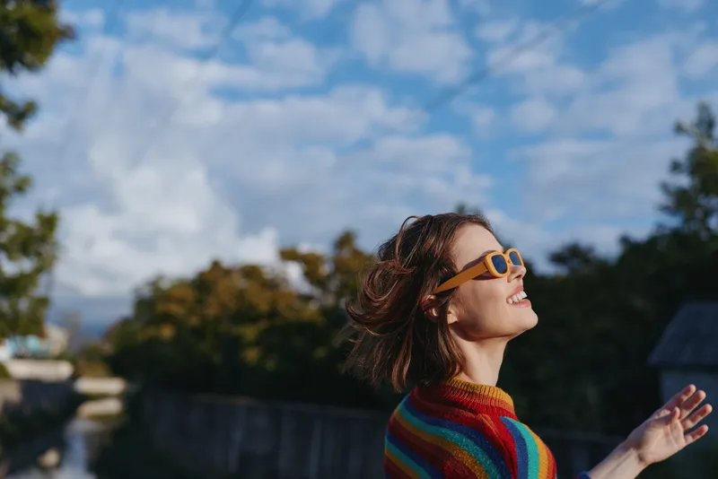 Young woman in her twenties wearing a colorful striped sweater and retro sunglasses, smiling with short hair outdoors by a river under blue sky, joyful travel portrait.