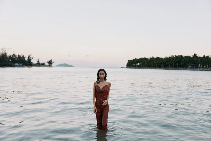 A woman stands knee deep in calm sea water at sunset, wearing a rust brown dress. Soft light reflects on the waves as distant trees frame a serene, minimalist fashion portrait.