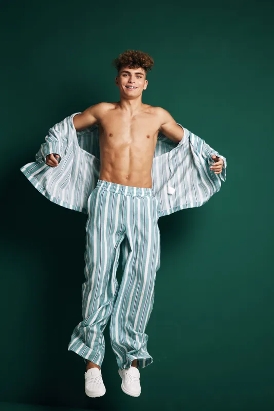 Young man jumping in striped pajamas with open shirt and white sneakers on green background, smiling and showing energetic mood in casual homewear outfit.