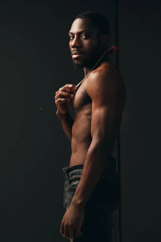 Creative conceptual portrait of a shirtless African man holding a red flower on his shoulder with serious expression, dark background, artistic visual style and strong emotional impact.