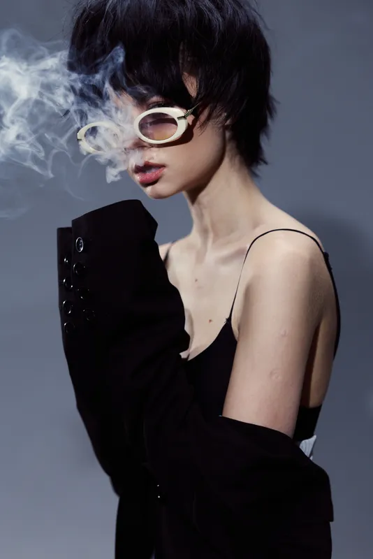 Fashionable woman with short hair smoking, wearing stylish sunglasses and black clothing in moody lighting.
