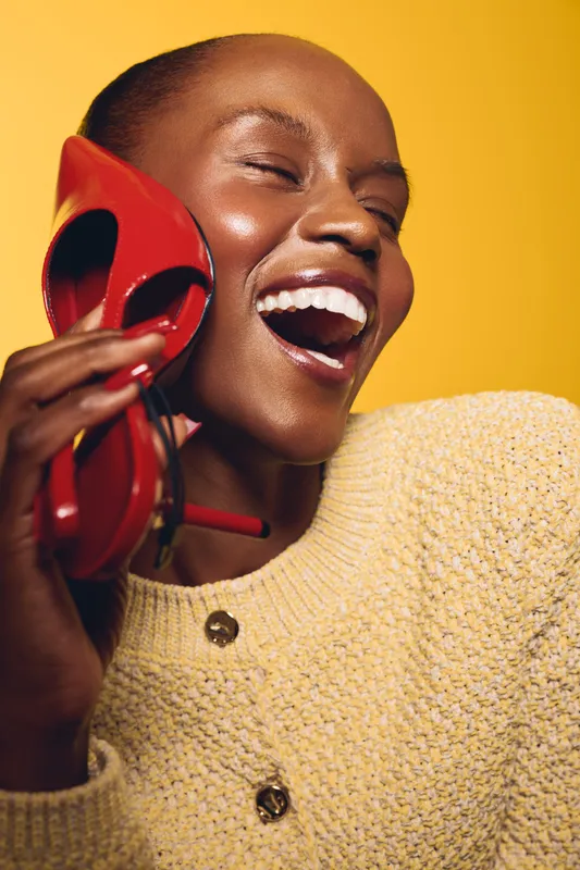 Happy dark skinned African woman with short hair laughs holding red high heel shoe near face. Yellow background and beige knitted sweater create warm joyful mood and fashion concept.