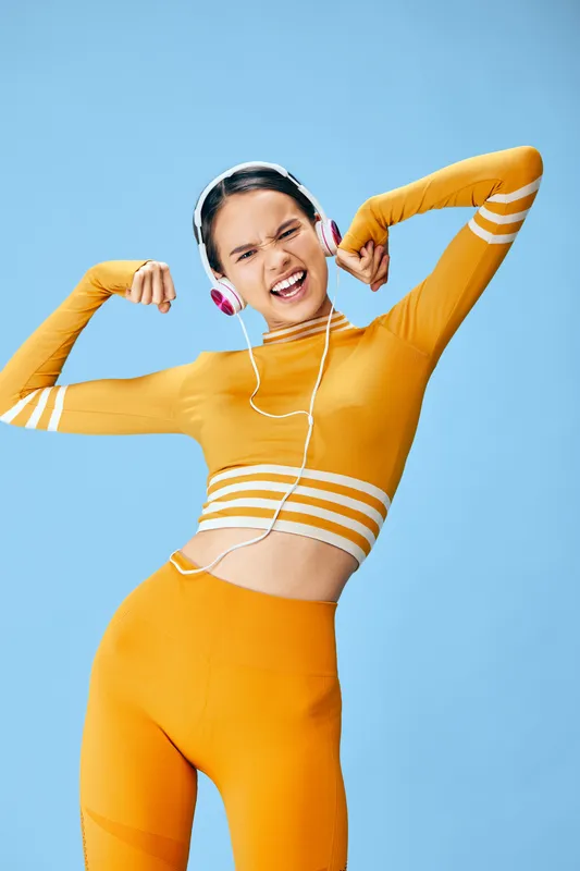 Fit young woman in bright orange activewear poses confidently with flexed arms wearing white headphones, upbeat fitness sport concept against smooth light blue studio background.