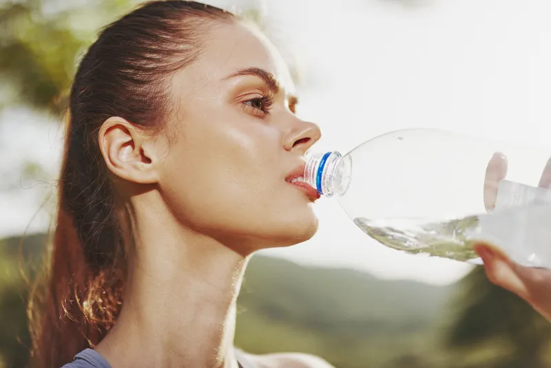 Young woman drinking water from a bottle in nature, healthy lifestyle, hydration, wellness, fitness, smiling, outdoor activity