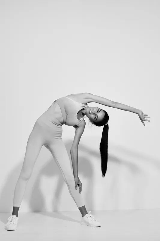 Black and white woman sport fitness stretching in activewear, demonstrating flexibility and balance with a focused expression against a plain backdrop.