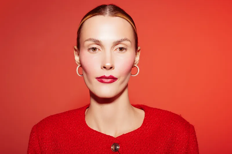 Portrait of confident woman with slicked hair and red lipstick wearing red sweater and hoop earrings on red background. Concept of fashion, beauty, and elegance in studio lighting.