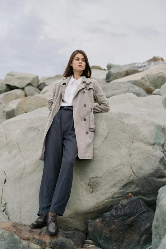 Young woman in grey coat and wide pants leans on large rock outdoors under cloudy sky. Casual fashion portrait with natural background and moody atmosphere.