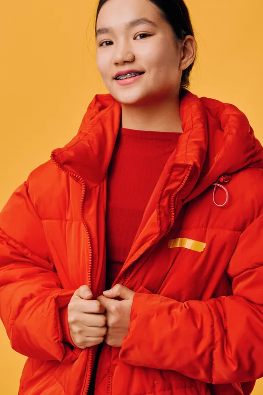 Young woman in red puffer jacket with zipper holding collar against orange background smiling confidently wearing red sweater, minimal makeup, and braces on teeth portrait in studio.