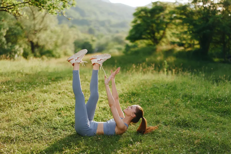 Young woman exercising outdoors in the summer, practicing leg raises in a lush green field, showcasing healthy lifestyle and fitness routine