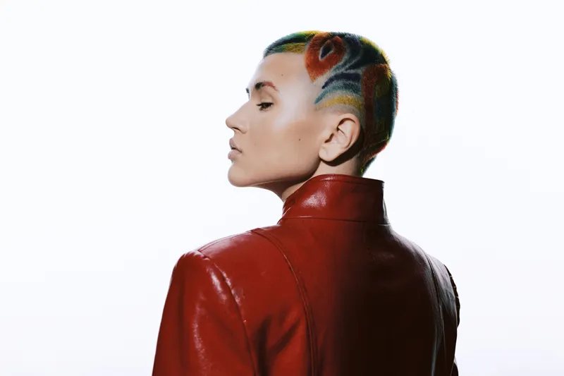 Woman with colorful painted short hair wearing a red leather jacket, posing confidently against a white background in a modern, artistic style.
