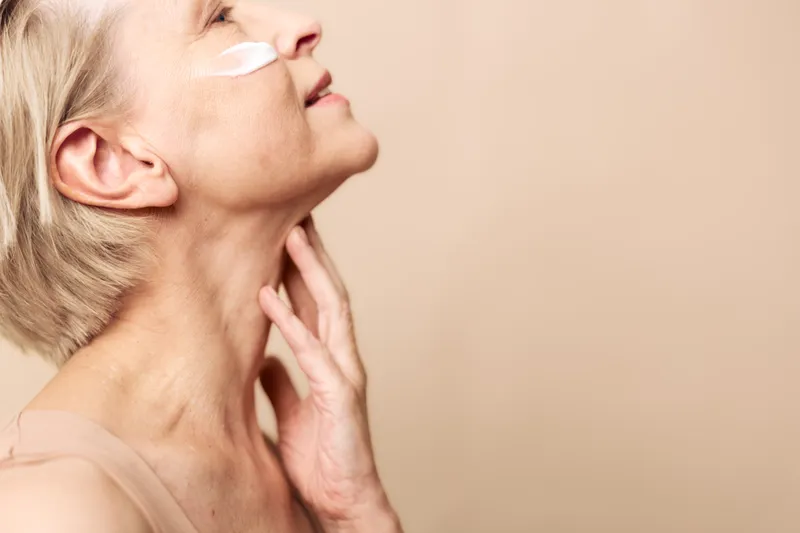 Mature woman with short blonde hair applies face cream on cheek, gently touching neck. Skincare concept for aging skin, beauty routine, and self-care on beige background.