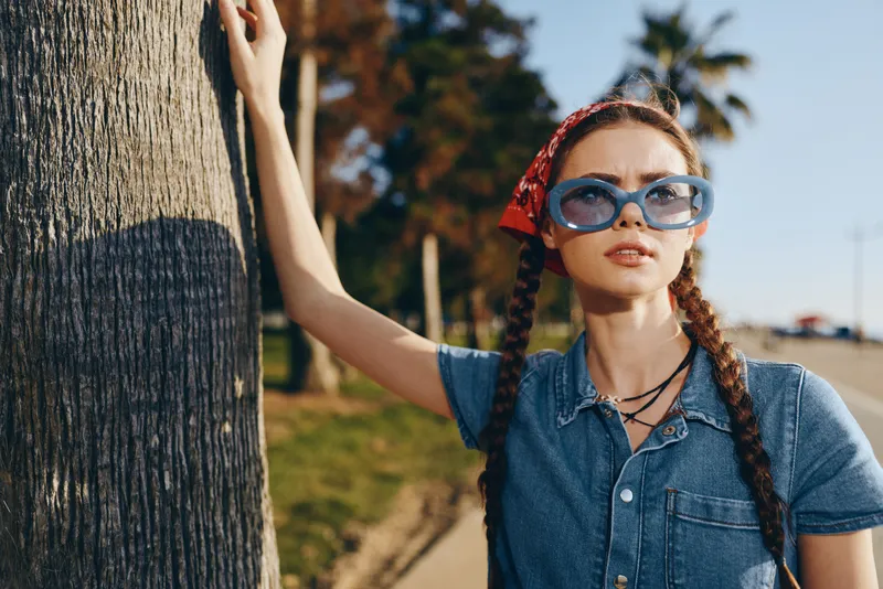 Lifestyle woman with modern boho-western denim street style wearing blue sunglasses and red bandana, captured in warm film color tones under natural sunlight.