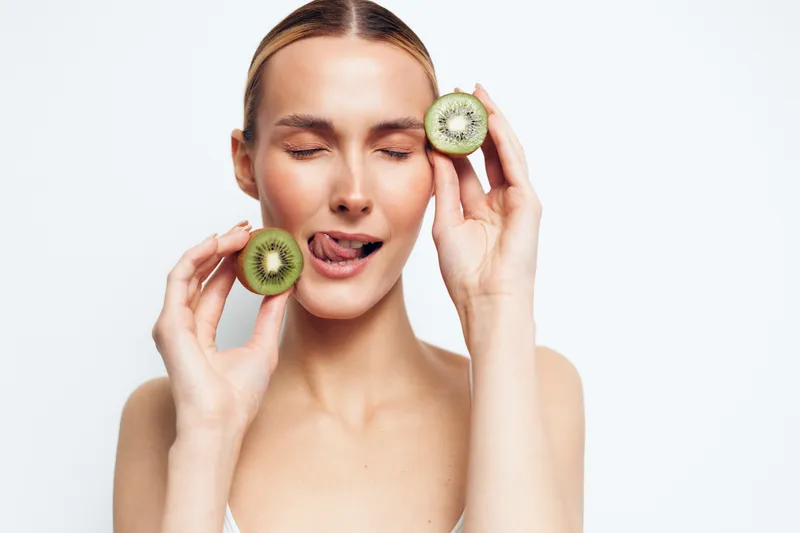 Smiling woman holds kiwi halves near her face, eyes closed, playful moment. Close-up on a light background. Fresh skincare concept, natural wellness, and self care. Soft lighting creates a warm mood.