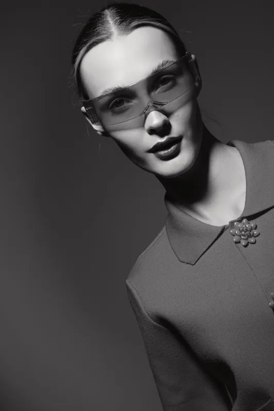Portrait of a young white woman with pale skin wearing sunglasses and a textured top with collar. Studio shot with dramatic lighting and monochrome effect.