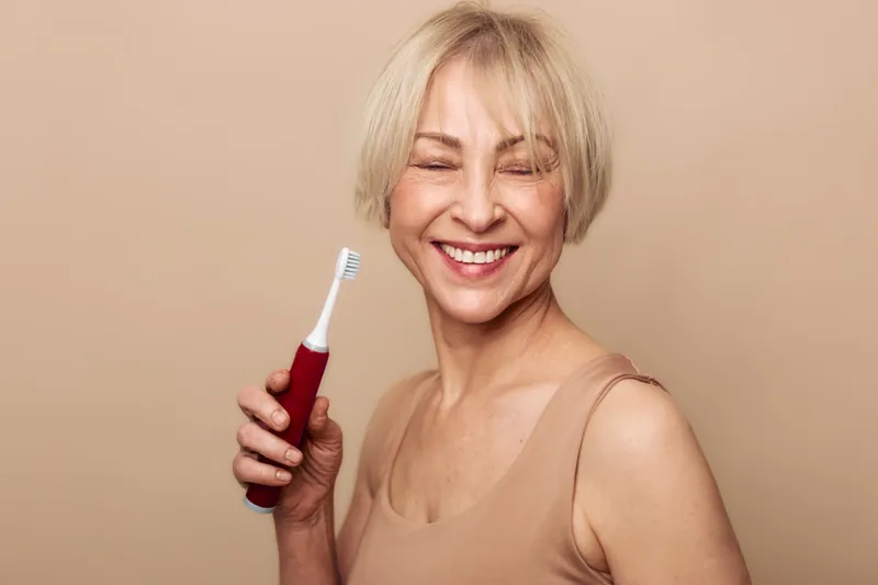 Smiling middle aged woman with short blonde hair holds electric toothbrush against beige background. Concept for dental care, oral hygiene, healthy lifestyle, and personal wellness.