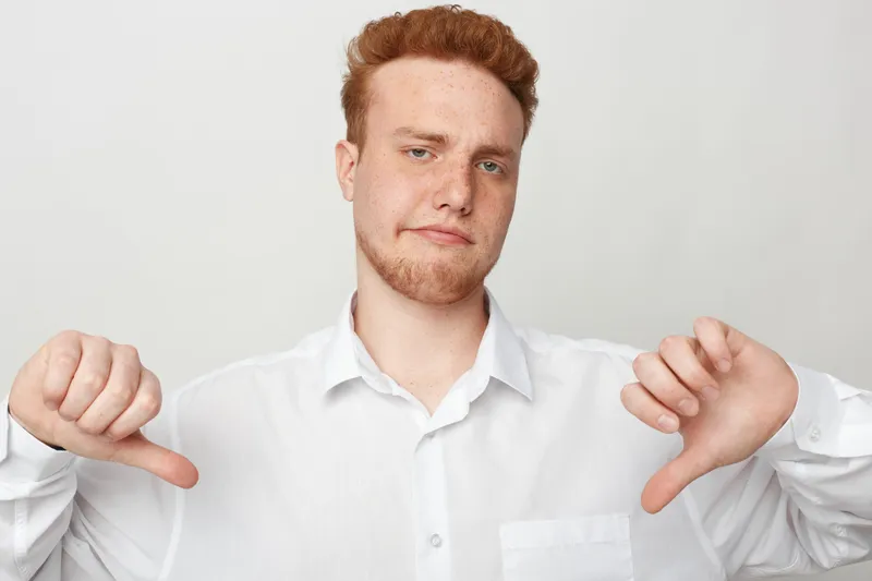Young man with red hair and beard wearing white shirt shows thumbs down gesture with both hands. Isolated on white background. Concept of disapproval, negative feedback, and dissatisfaction.