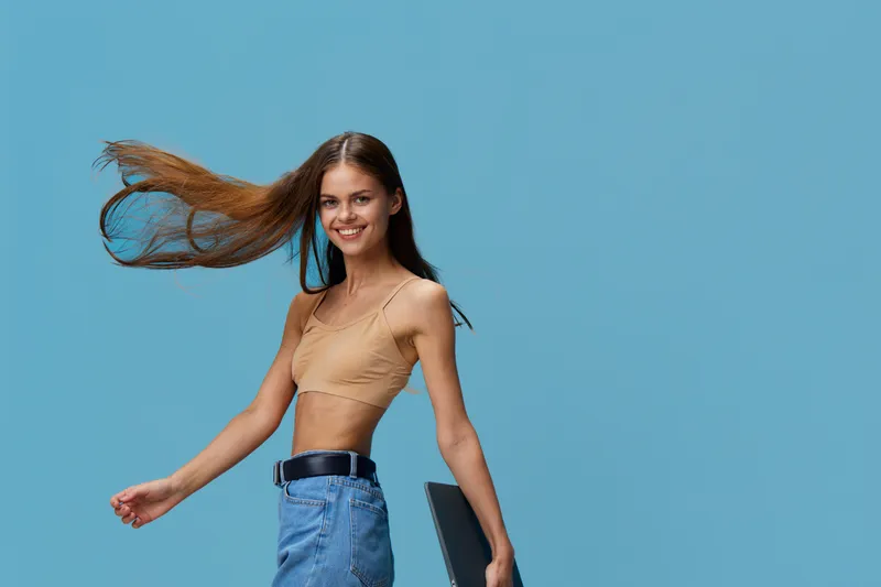 smiling woman with long hair wearing casual beige top and denim skirt stands against blue background holding laptop, hair flowing, youthful and carefree expression