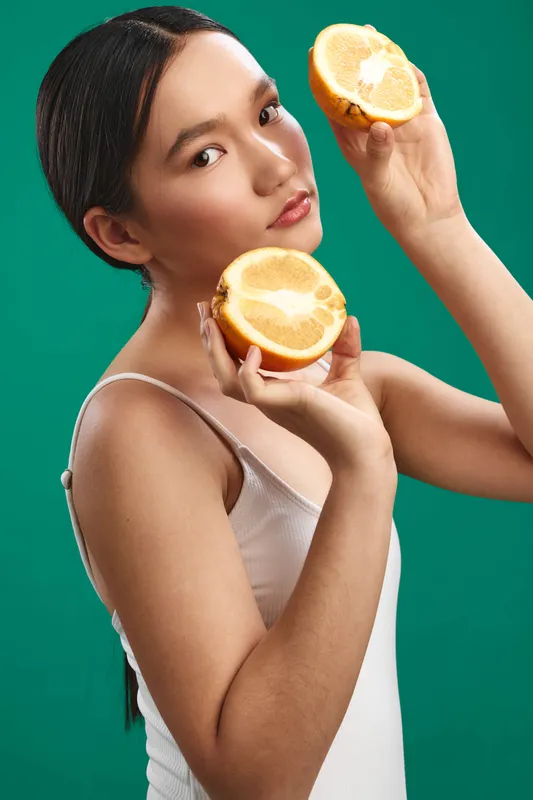 Asian woman holds fresh orange halves against a vibrant green backdrop, presenting a bright, refreshing still life that highlights healthy snacking and bold color.