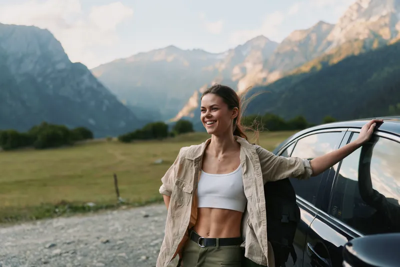 Smiling woman in casual clothes leaning on car door in mountain landscape. Outdoor lifestyle, travel, freedom, and adventure concept with natural light and relaxed mood.